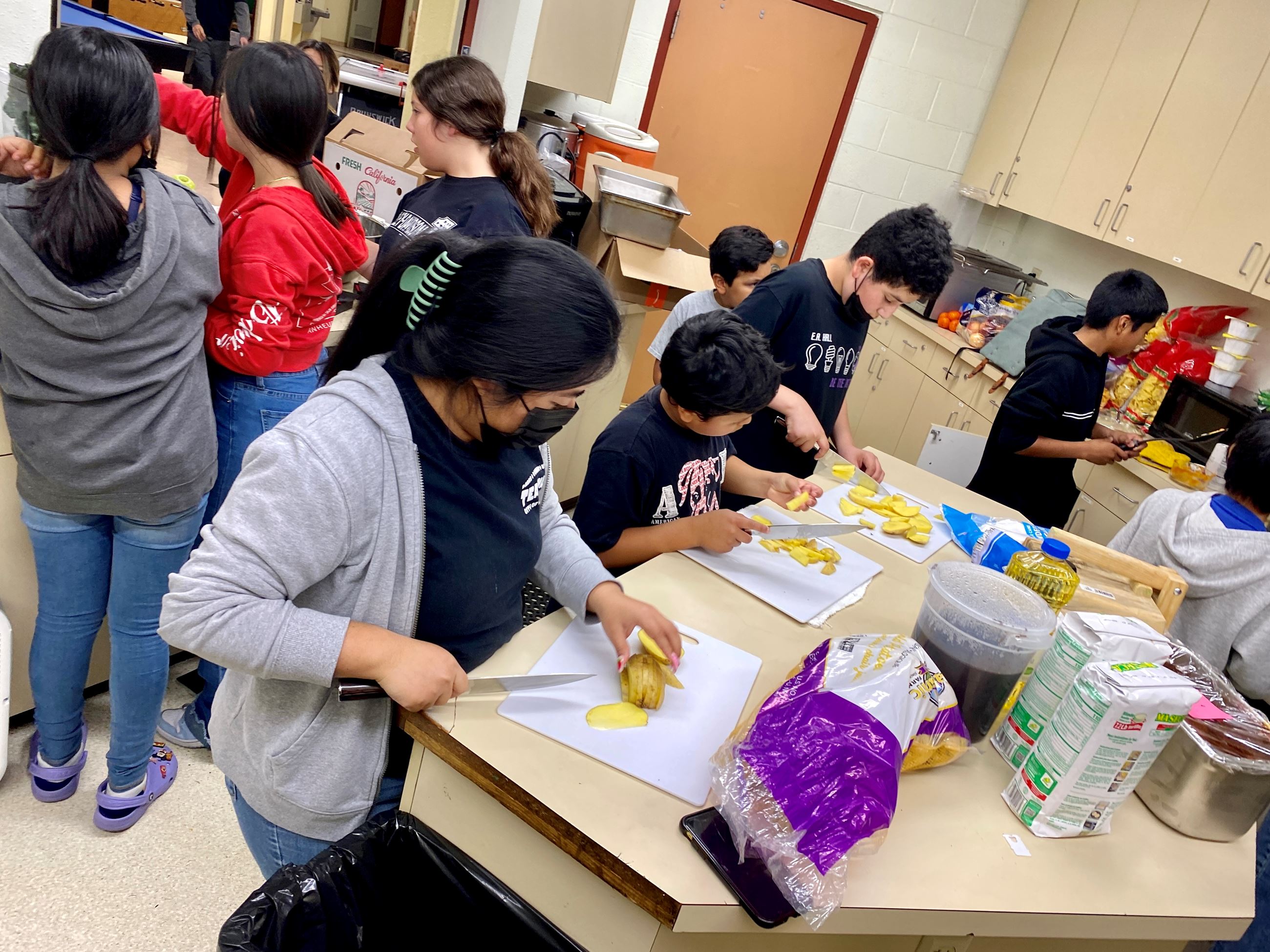many teens in a kitchen cooking, three teens cutting potatoes